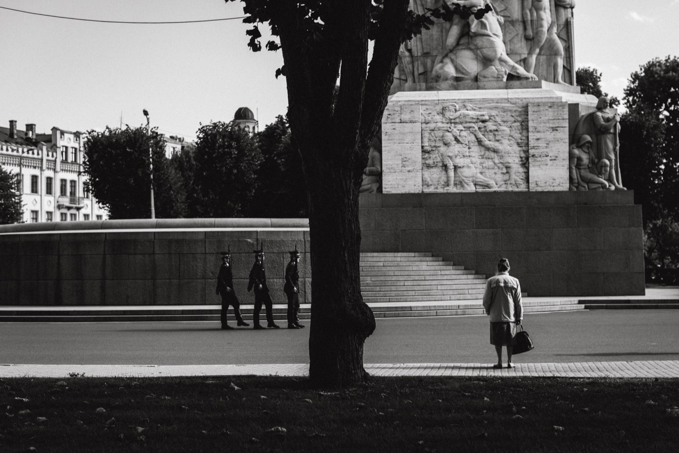 The Freedom Monument in central Riga — three soldiers march in formation; a woman in the foreground watches them.