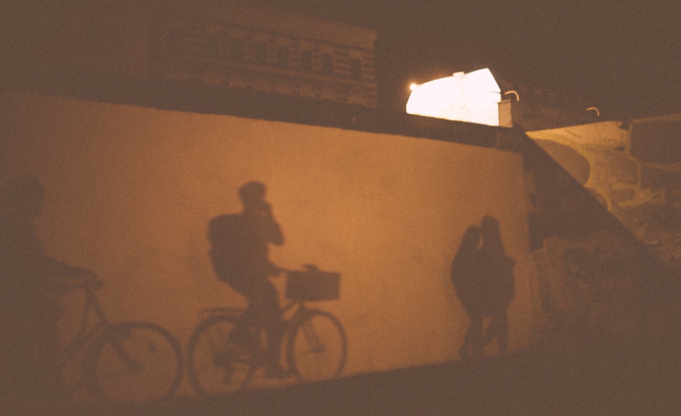 Two cyclists and a couple pass as silhouettes against the interior face of the East Side Gallery at night.