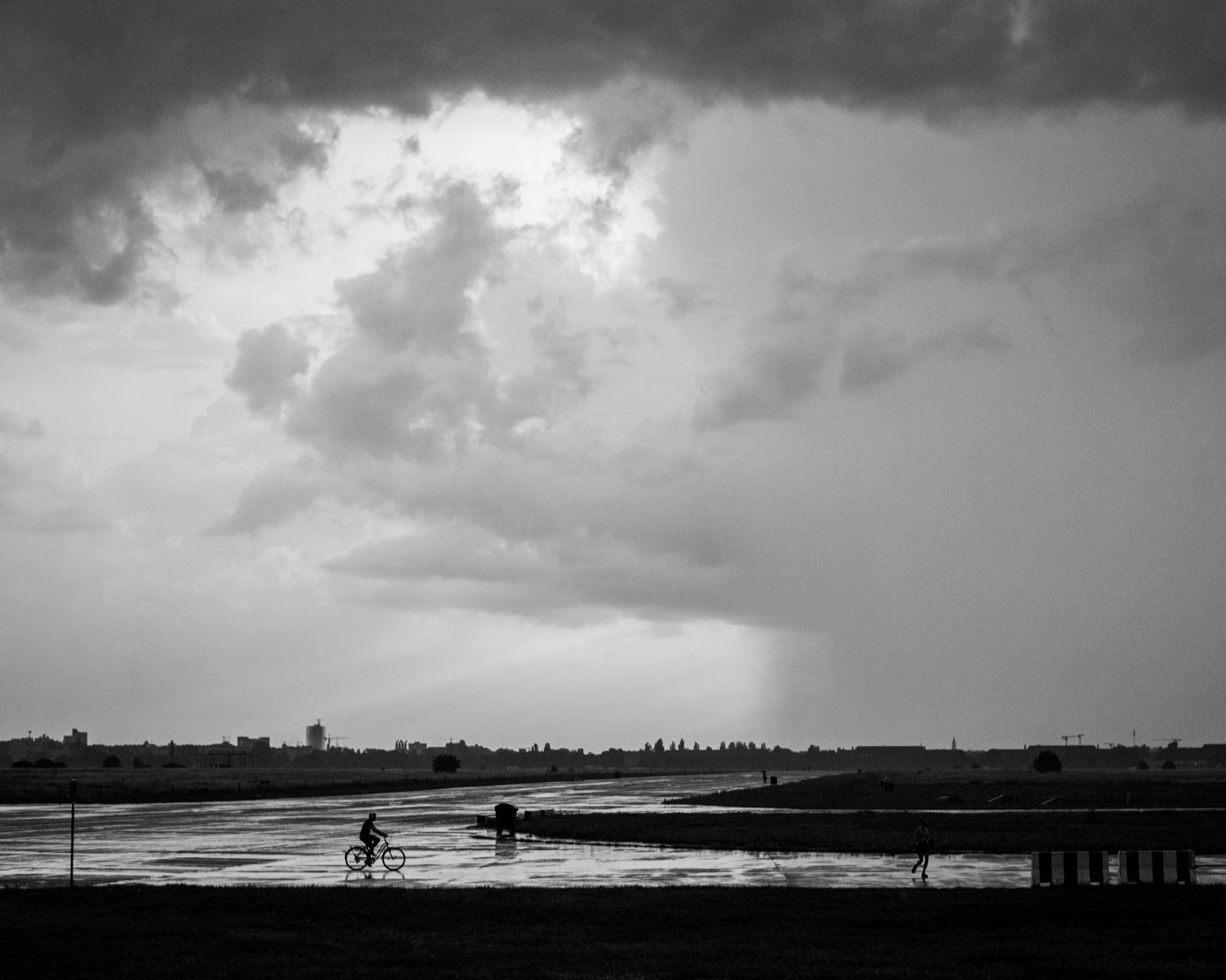 A black-and-white frame from Tempelhof after rain &mdash; a bicyclist crosses the frame against rain-bright distance, the slick runway reflecting the sky.