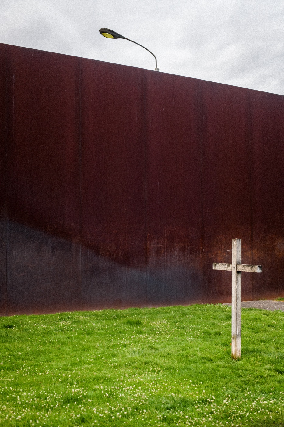A short stretch of corrugated metal wall near Bernauer Stra&szlig;e, with a streetlight and a small wooden cross in the grass.