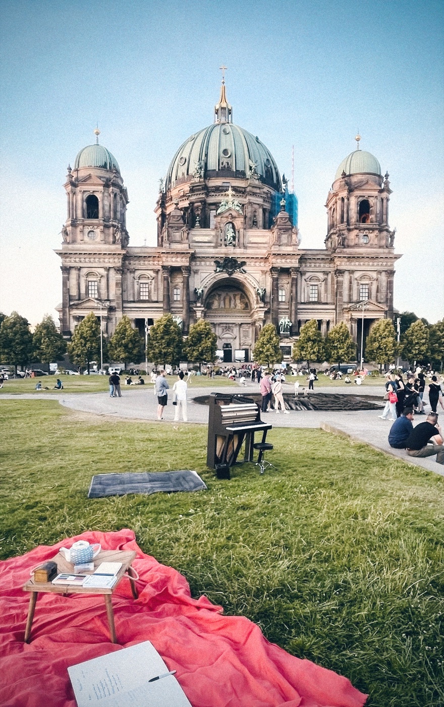 A red picnic blanket on the Lustgarten plaza with a keybird set up; the Berliner Dom rises monumentally behind.